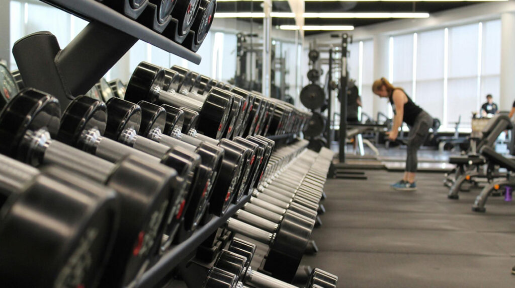 A woman in a gym standing in front of a rack of dumbbells.