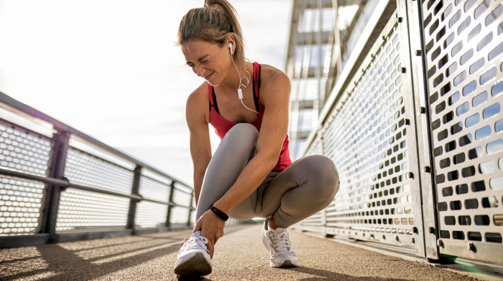 While on a run, a woman crouches down and grimaces as she holds her injured ankle in pain.