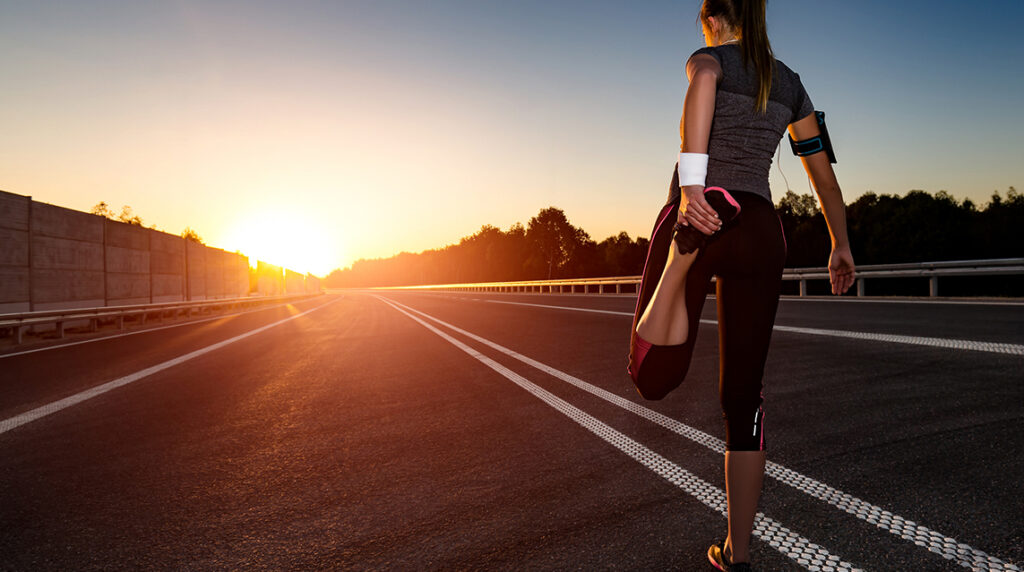 A woman stretches her quads as she stands on a track facing the sunrise, ready to start her half marathon training schedule for beginners
