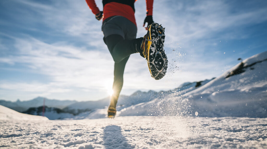 A male trail runner runs along a snow-covered mountain trail with glaciers and distant snowy peaks in the background.