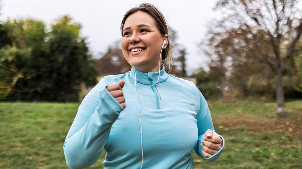 A smiling woman outdoors wearing a blue athletic jacket and wired headphones.