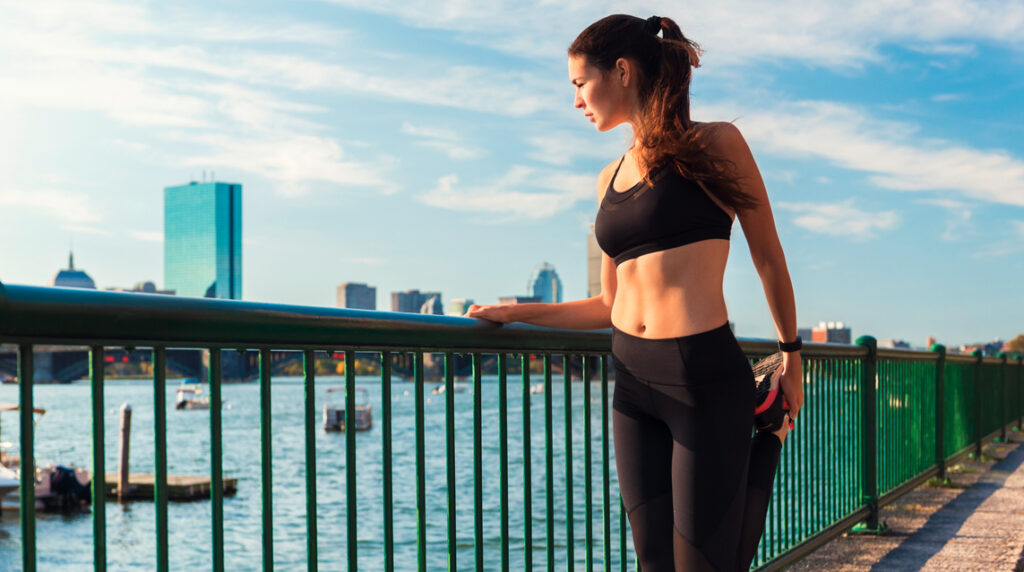 A woman stretches in preparation for a meeting with a Boston running club while looking at a waterfront view of the city’s skyline.