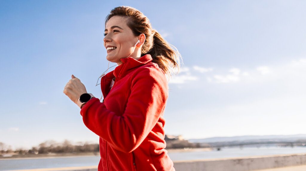 A woman in proper running form jogging by the waterfront with earbuds, smiling.